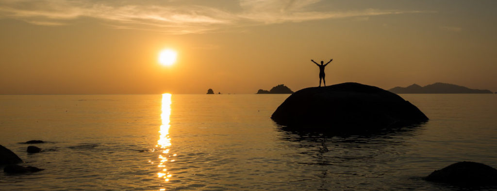 Photo of victorious person standing on a rock just offshore at sunset
